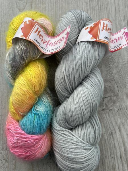 Zwei Stränge handgefärbte Wolle: links Herzfasern No. 50 „Ohne Kommentar“ in leuchtendem Gelb, Pink, Türkis und Grau mit Mohairstruktur, rechts Herzfasern No. 11 „Silber White“ in hellem Silbergrau auf glatter Merino-Basis. Fotografiert auf hellem Holzunt